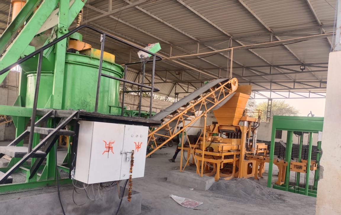 Green pan mixer feeding a yellow block-making press by conveyor, under a production shed
